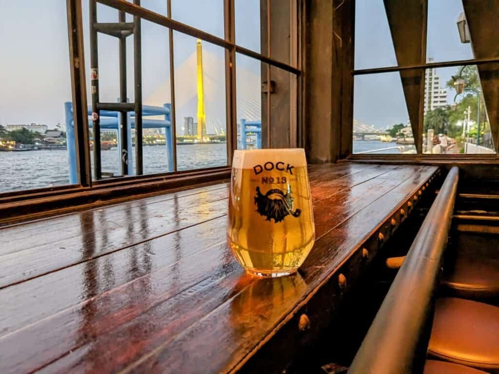 Glass of beer on a wooden table with water in the background and the sunset on a bridge. At Sheepshank Public house or Dock No. 13 in Bangkok Thailand.