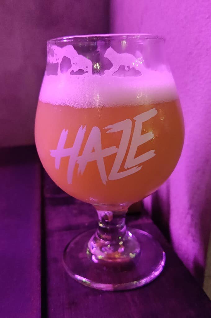 Cloudy IPA craft beer in a Haze branded goblet style glass on a wooden table next to stone walls in Bangkok, Thailand.